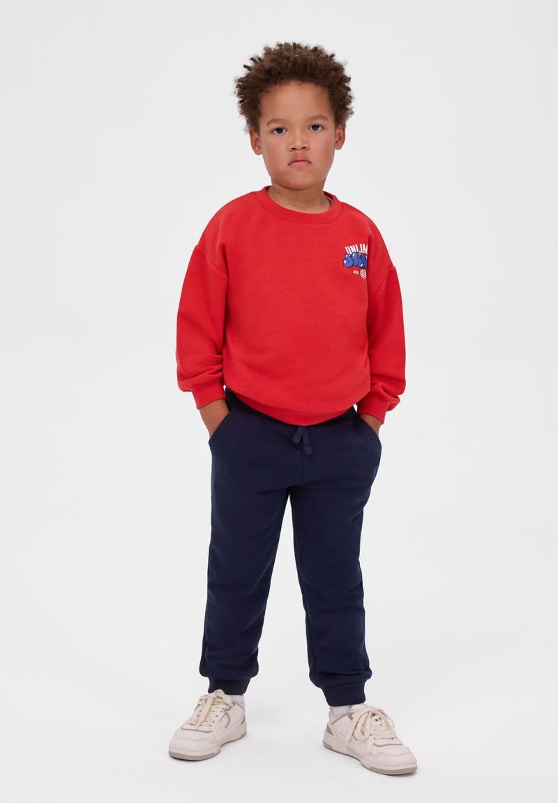 Jeune enfant aux cheveux bouclés, debout, mains dans les poches, portant un sweat-shirt rouge, un pantalon bleu foncé et des baskets blanches sur un fond uni.