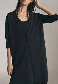 Femme portant un long cardigan noir ample sur un haut noir, un bras levé et les doigts touchant ses cheveux.
