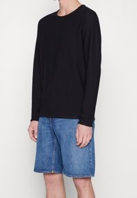 Schwarzes Langarmshirt aus weichem Stoff mit rundem Halsausschnitt. Kombiniert mit hellblauen Jeansshorts mit geradem Schnitt und Vorderaschen.