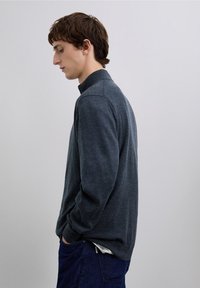 Dunkelgrauer Pullover mit hohem Kragen und langen Ärmeln, aus einem strukturierten Strickstoff gefertigt. Trägt man über blauen Jeans und zeigt eine legere Passform.