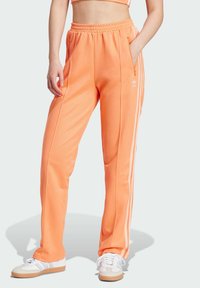 Orange sweatpants with white side stripes, elastic waistband, and pockets, paired with white sneakers. Smooth fabric and relaxed fit.