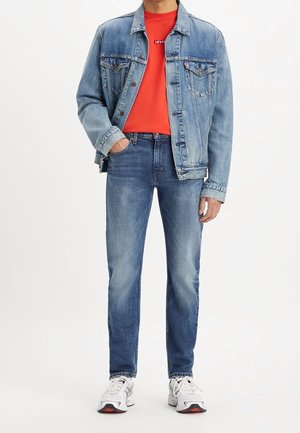 Person trägt blaue Jeansjacke und Jeans, rotes Levi's T-Shirt, weiße Turnschuhe, steht mit einer Hand in der Tasche vor weißem Hintergrund.