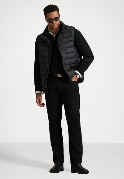 Gilet matelassé noir avec un col montant sur une chemise à manches longues noire, associé à un pantalon noir et des bottes en cuir. Il présente une montre en argent.