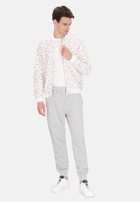 Lichtgewicht bomberjack met een kleurrijk bloemenpatroon op een witte achtergrond; gecombineerd met grijze joggingsbroek en witte sneakers.
