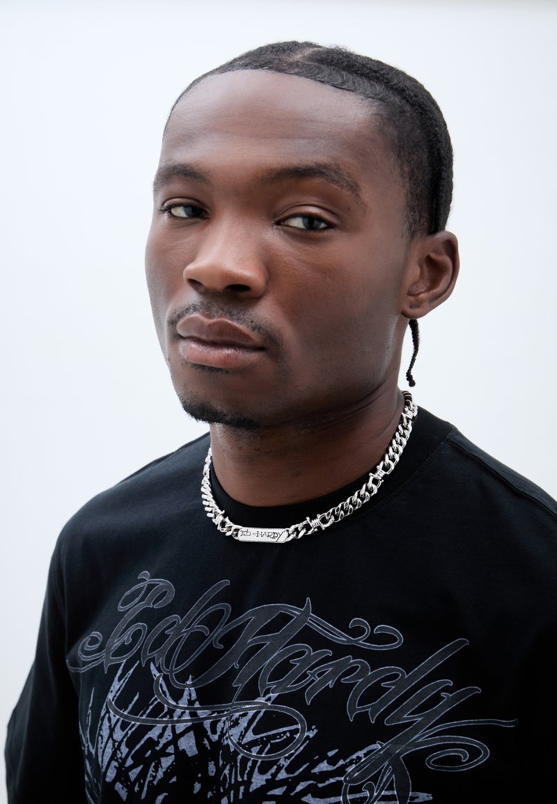 Jeune homme aux cheveux tressés portant un t-shirt noir Ed Hardy et une chaîne argentée assortie, regardant calmement vers la caméra.