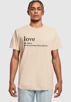 T-shirt en coton beige avec texte imprimé noir : "amour (lat. lubère) un sentiment intense d'affection profonde." Manches courtes, coupe décontractée.