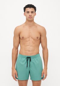 Turkise badeshorts med svart snøring, elastisk linning og et glatt, lett stoff. Modellen står vendt mot kamera.