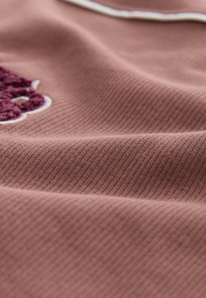 Tekstiilikangas pölyisen ruusun sävyssä, jossa on poimutettu pinta. Siinä on tumman viininpunainen kirjailtumuotoilu ja valkoinen korostus.