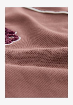 Tissu textile de couleur rose poudré avec une texture côtelée. Présente un motif brodé en bordeaux foncé avec un contour accentué blanc.