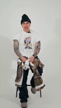 Ung mann med tatoveringer som har på seg en lue, hvit t-skjorte med trykk, svarte bukser, og holder en kamuflasjejakke mens han sitter på en krakk.