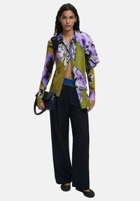 Camicia a bottoni floreale con tonalità viola e verde, abbinata a pantaloni neri a gamba larga. Il modello tiene una borsa nera con una tracolla intrecciata.