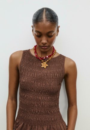Top senza maniche color marrone con superficie testurizzata, abbinato a una collana di perline multicolore con pendente a forma di fiore.