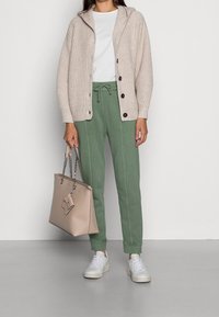Cárdigan de punto Beige con capucha, pantalones de chándal verdes con cordón, camiseta blanca, zapatillas claras y un bolso beige pálido con correas de cadena.