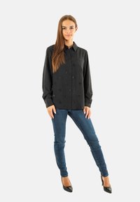 Camicia nera a maniche lunghe con un motivo floreale testurizzato, colletto a punta, abbinata a jeans blu aderenti e tacchi neri.