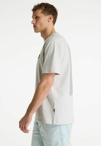 Joven con camiseta gris claro y vaqueros azul claro de pie de lado contra un fondo blanco liso.