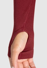 Rød ribbet langærmet bluse med en tommelfingerhuludskæring, der har en glat tekstur og tætsiddende design, som fremhæver den åbne hånd.