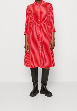 Kvinde iført en rød og hvid vertikalt striped knap-op kjole med taljebånd og sorte knæhøje chunky støvler, stående på en ensfarvet baggrund.