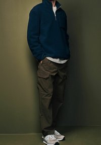 Marineblauer Fleece-Viertelreißverschluss-Pullover mit einer Brusttasche, über einem gestreiften Hemd getragen, kombiniert mit olivgrünen Cargo-Hosen und grauen Sneakers.