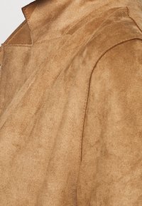 Sämja ytterplagg i varm tan-färg, med en mjuk textur, skarp krage och rena sömmar. Tyget verkar något borstat.