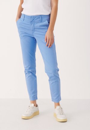 Persoon die lichtblauwe slim-fit broek en witte sneakers draagt, staand met één hand in de zak tegen een effen achtergrond.