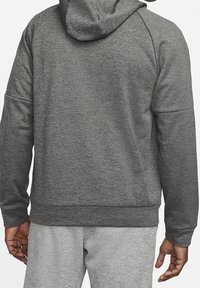 Personne portant un sweat à capuche gris foncé et un pantalon gris clair, vue de dos avec les bras détendus le long du corps.