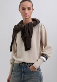 Mujer con suéter de mangas abullonadas beige, suéter marrón sobre los hombros, jeans azules y dos pulseras gruesas en la muñeca izquierda.