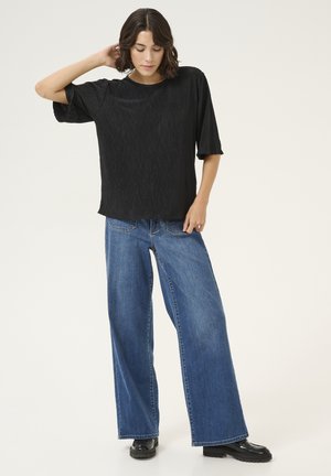 Black pleated short-sleeve top paired with wide-leg blue jeans. Model wears black chunky boots. Simple and relaxed fit design.