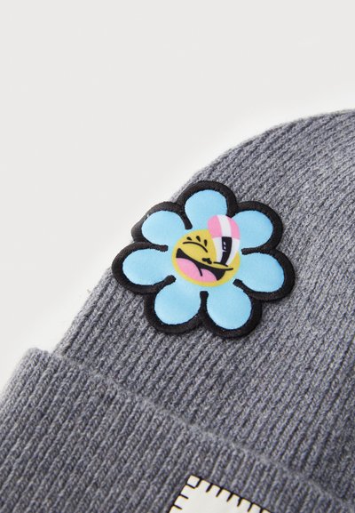 Bonnet tricoté gris avec une fleur bleue brodée représentant un visage souriant en cartoon et des contours noirs. Tissu texturé avec un motif en côtes.