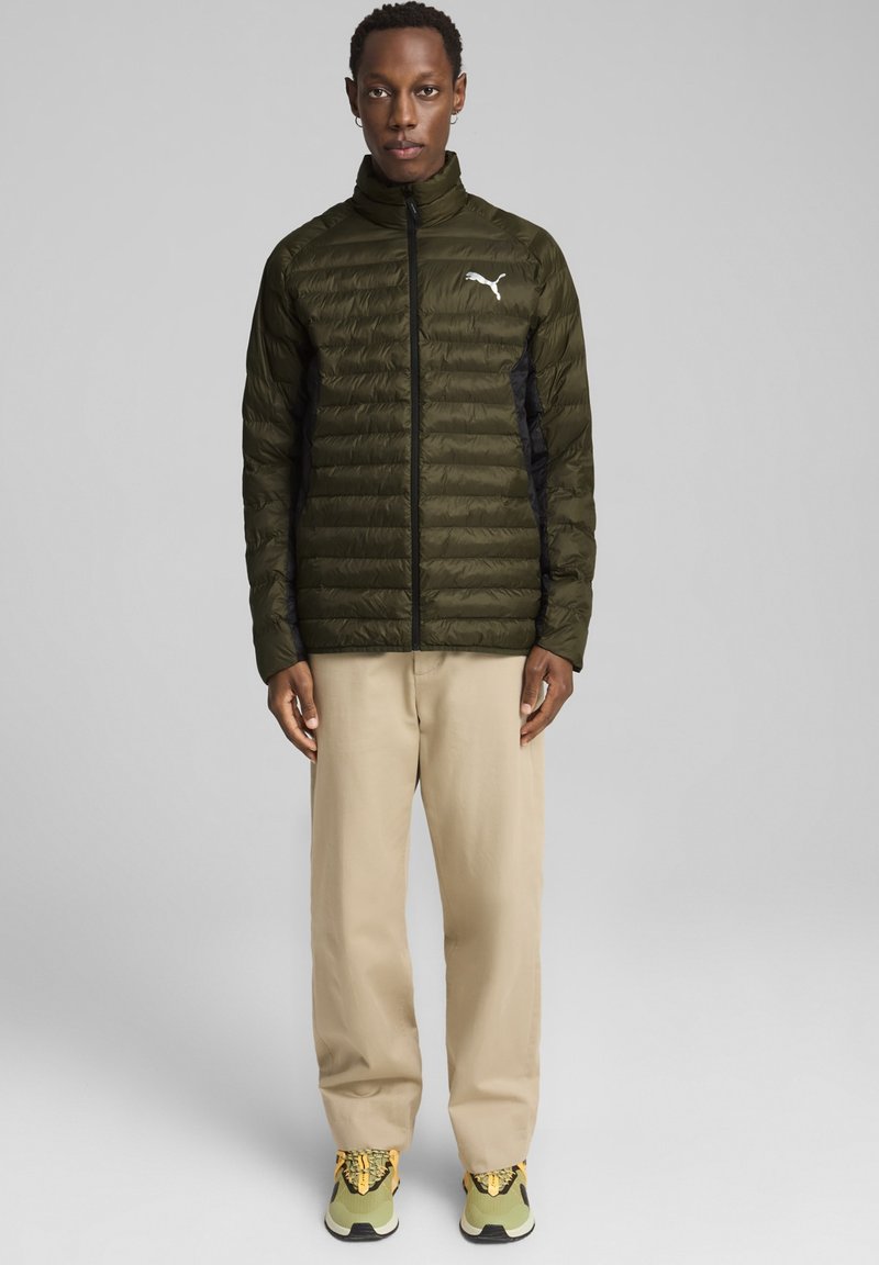 Puffy jakke i olivengrøn med sorte sidepaneler, lynlås lukning, høj krave og et hvidt logo. Båret med beige bukser og grønne sneakers.