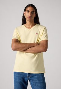 Tricou galben cu guler în V, fabricat dintr-un material moale, având un mic logo roșu Levi's pe pieptul stâng, asociat cu blugi din denim.