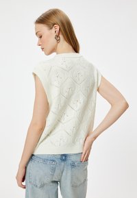 Dzianinowy bezr�ękawnik w kolorze off-white z heksagonalnym wzorem teksturowym i prążkowanymi krawędziami. Połączony z jasnoniebieskimi jeansami o wysokim stanie.