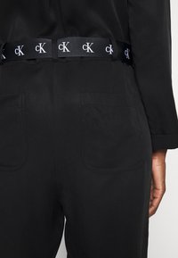 Ceinture en tissu noir comportant le logo "cK" répété en blanc. Associée à un pantalon noir avec deux poches arrière, des coutures visibles et une texture lisse.