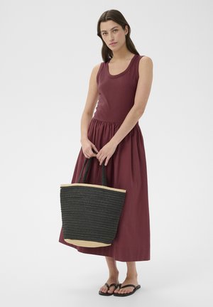 Femme portant une robe bordeaux sans manches et des sandales noires, tenant un grand sac fourre-tout tissé noir avec une bordure beige sur un fond uni.