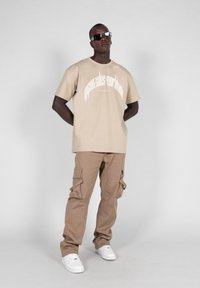 Beige grafisk t-skjorte med hvit tekst, kombinert med kaki cargo-bukser med sidelommer. Modellen har på seg solbriller og hvite joggesko.