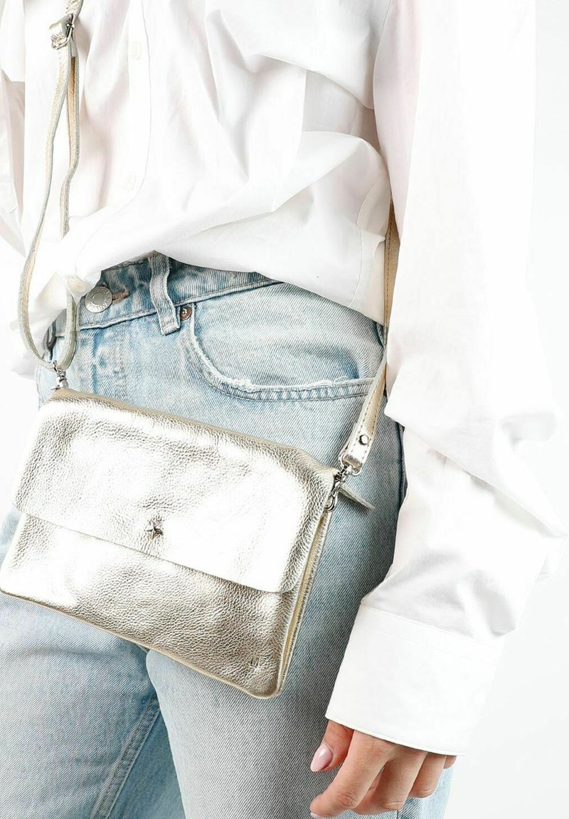 Metalen zilveren crossbody tas met een gestructureerd oppervlak, uitgerust met een vlakke klep sluiting en verstelbare schouderriem, gedragen met lichte denim.