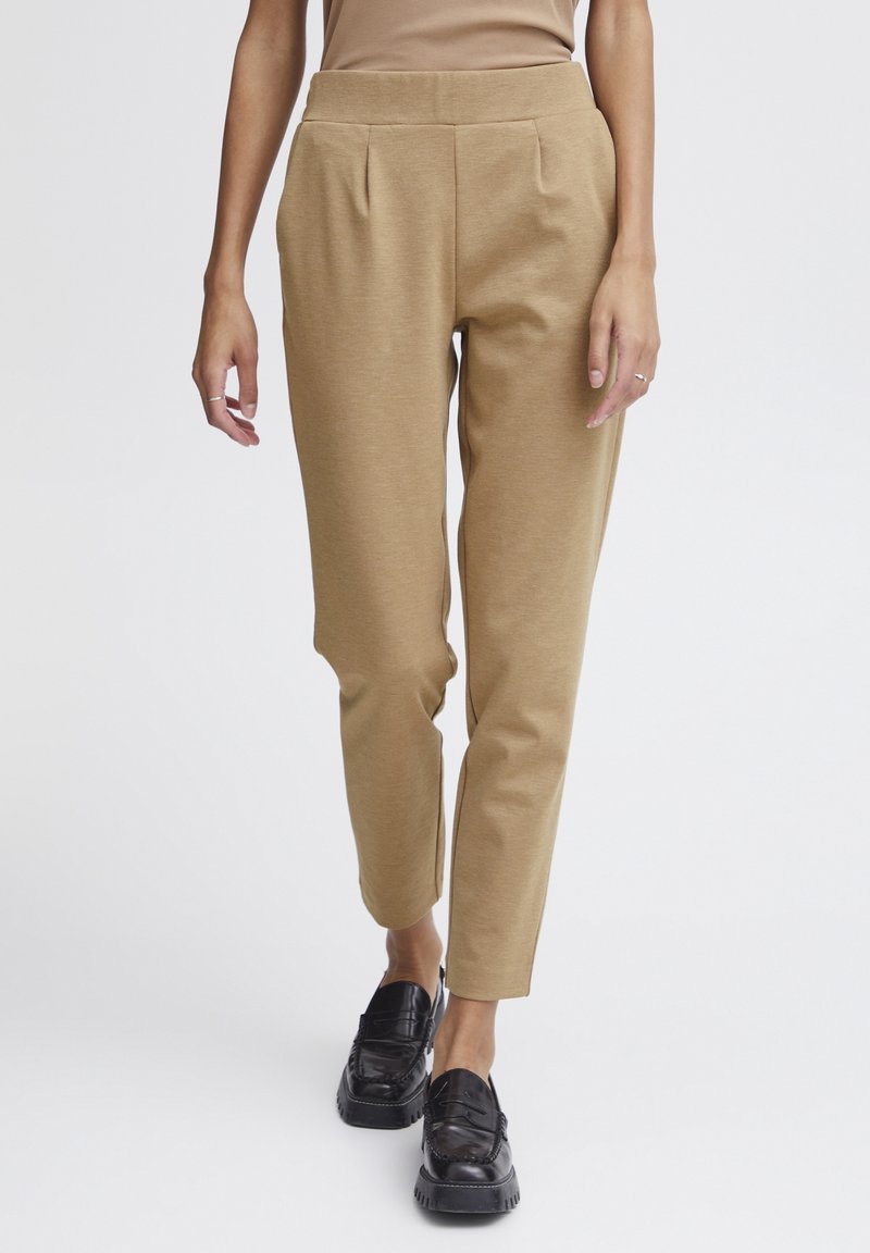 Femme portant un pantalon beige fuselé et des mocassins noirs, debout devant un fond clair uni.