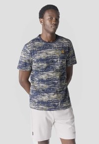 Junger Mann mit kurzen lockigen Haaren, der ein blau gemustertes T-Shirt und hellfarbene, strukturierte Shorts trägt und vor einem schlichten Hintergrund steht.