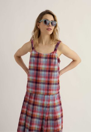 Nő kék napszemüvegben, hozzáillő piros, kék és narancssárga kockás ujjatlan felsőben és szoknyában, kezei a csípőjén pihennek.
