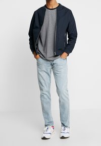 Marinebomberjakke, gråstribet T-shirt, lyseblå jeans og hvide sneakers med blå og røde accenter. Glat tekstur, afslappet design.