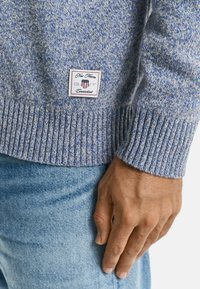 Hellesblau gestrickter Pullover mit geripptem Saum. Ein kleines Markenschild an der Seite zeigt "New Haven, GANT, Connecticut".