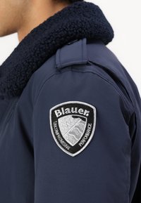 Jūras zilas krāsas jaka ar teksturētu vilnas apkakli, aprīkota ar kreiso plecu uzšuvumu, uz kura attēlots "Blauer" logo sudraba un melnā krāsā.