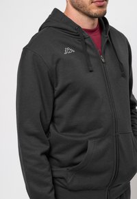 Sötét szürke cipzáras kapucnis pulcsi puha anyagból, elülső zsebbel és kis logóval a bal mellen. Állítható zsinóros kapucni.