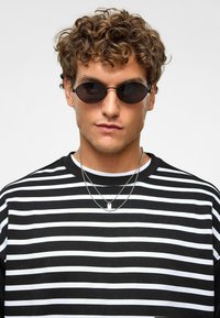 Ronde zwarte zonnebril met dunne monturen, gedragen met een zwart-wit gestreepte lange mouwen shirt. Heeft twee zilveren kettingen om de nek.