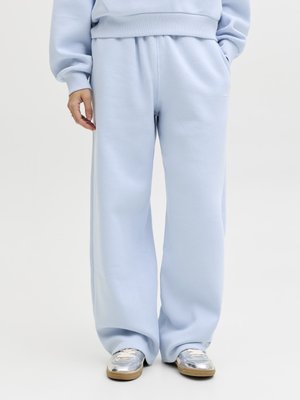 Persona che indossa pantaloni della tuta larghi azzurri chiaro, felpa coordinata e sneaker metallizzate argento su sfondo neutro.