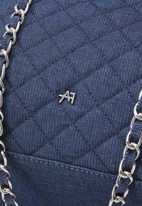 Granatowa torba na ramię z pikowanej tkaniny z srebrnym łańcuszkiem i małym metalowym detalem z logo. Tekstura jest gładka z wzorem wypukłych kwadratów.