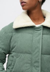 Casaco acolchoado verde com nervuras e gola de pêlo falso creme. Apresenta fecho frontal e bolso lateral. Tecido texturizado que realça o interesse visual.