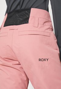 Rosa Skihosen mit einem strukturierten schwarzen Bund. Mit schwarzem "ROXY"-Logo, zwei Gesäßtaschen und glatter Stoffstruktur.