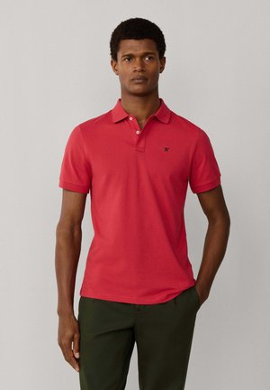 Man staat en draagt een rood poloshirt met korte mouwen met een klein zwart logo en donkergroene broek, één hand in de zak.