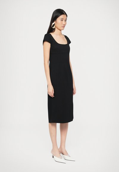 Melnā pieguļošā kleita ar kvadrātveida izgriezumu un īsām piedurknēm, izgatavota no gludas auduma. Stilizēta ar baltām ar smailu purngalu mūlēm un zelta riņķveida auskariem.