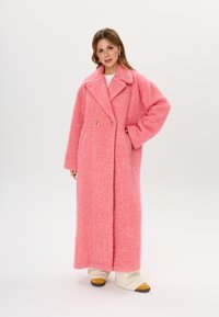 Langer, flauschiger Mantel in leuchtendem Pink mit Reverskragen, doppelt gereihtem goldenen Knöpfen und Seitentaschen. Trägt er über weißen und cremefarbenen Schichten.